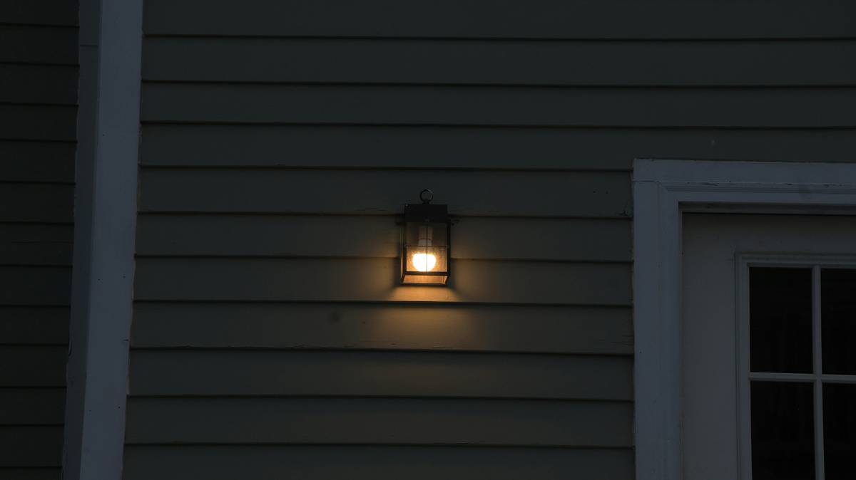 Outdoor illumination seasonal flood light home mounted on a cedar pergola with string lights and autumn pumpkins visible in foreground